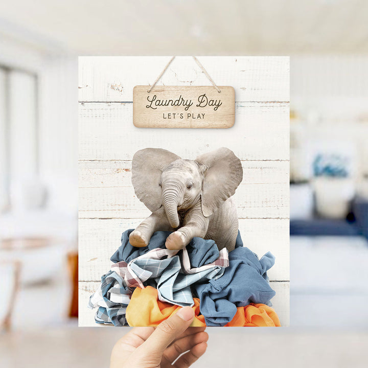 A hand holding a sign that says "Laundry Day Let's Play" with an image of a baby elephant sitting on a pile of clothes.