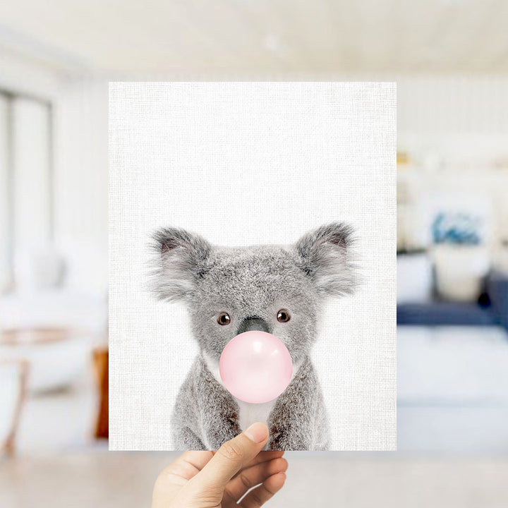 A hand is holding up a picture of a koala bear blowing a bubble with a pink bubble gum bubble.