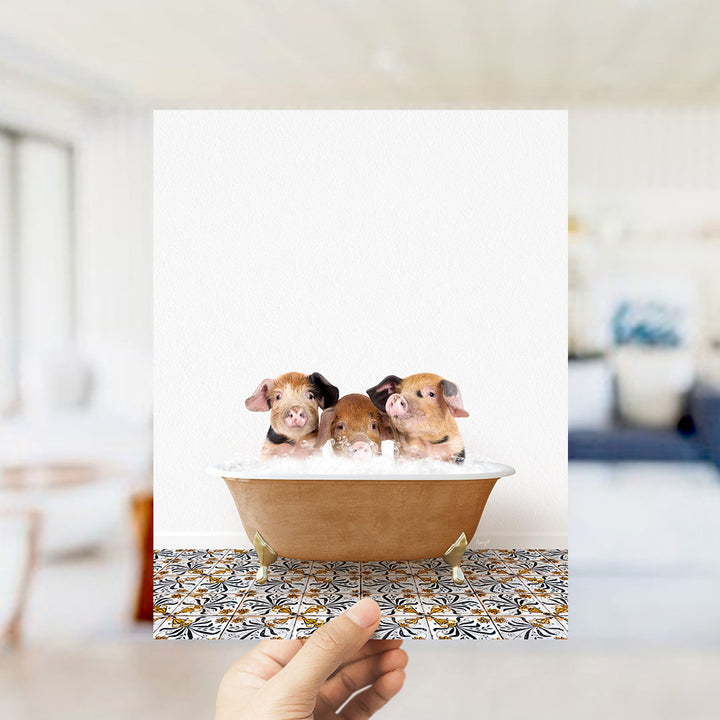 A hand holds up a framed image of three adorable pigs sitting in a bathtub filled with bubbles.