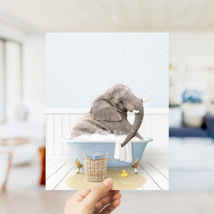 A hand holds up a framed image depicting an elephant sitting in a bathtub filled with bubbles, surrounded by a yellow rubber duck and a white towel.