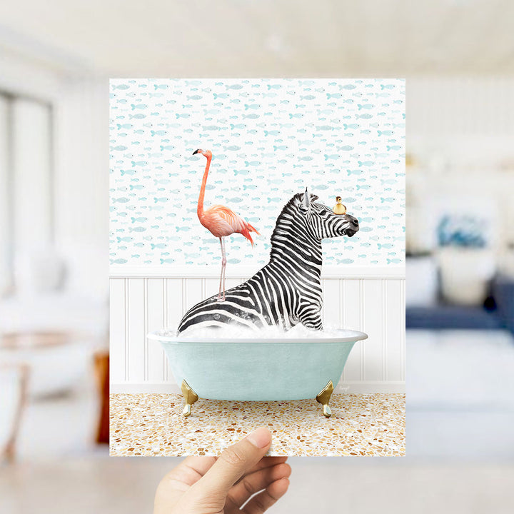 A hand holds up a framed image depicting a zebra sitting in a bathtub, with a pink flamingo standing on its back.