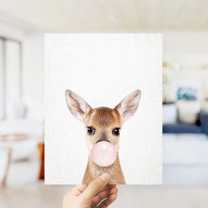 A hand is holding up a white square with a picture of a deer blowing a bubble with a pink bubble gum bubble.