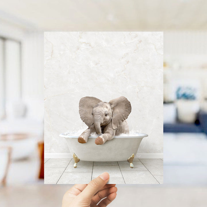 A hand holds up a framed image of a baby elephant sitting in a bathtub filled with water, with its large ears spread out.