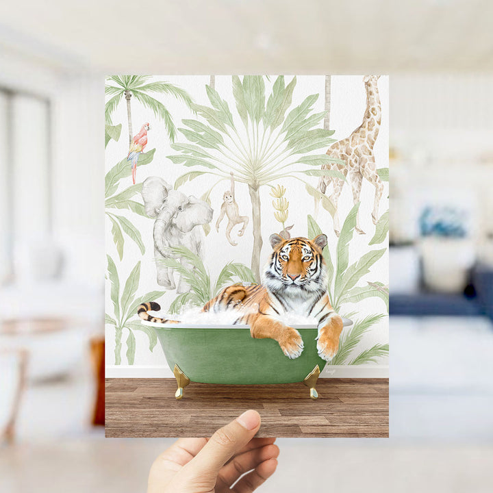 A hand holds up a framed print featuring a tiger lying in a bathtub surrounded by tropical foliage, including a giraffe, elephant, and parrot.