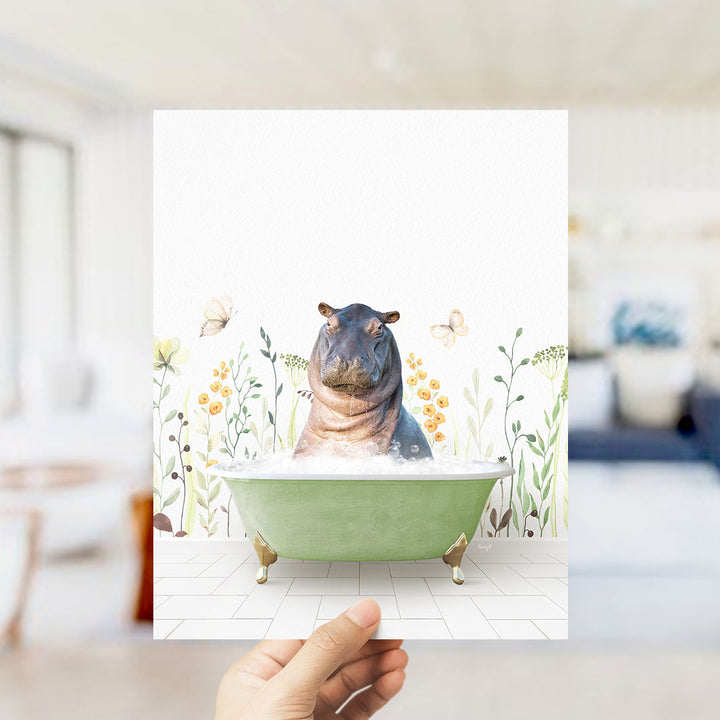 A hand holds up a framed image depicting a hippopotamus sitting in a green bathtub surrounded by flowers and butterflies.