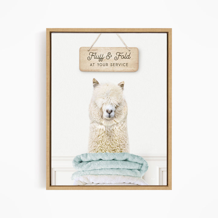 A fluffy white alpaca sits atop a stack of light blue towels, with a wooden sign above that reads "Fluffy & Fold at your service".