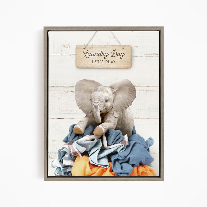 A framed image of a baby elephant sitting atop a pile of laundry, with a sign above that says "Laundry Day Let's Play".