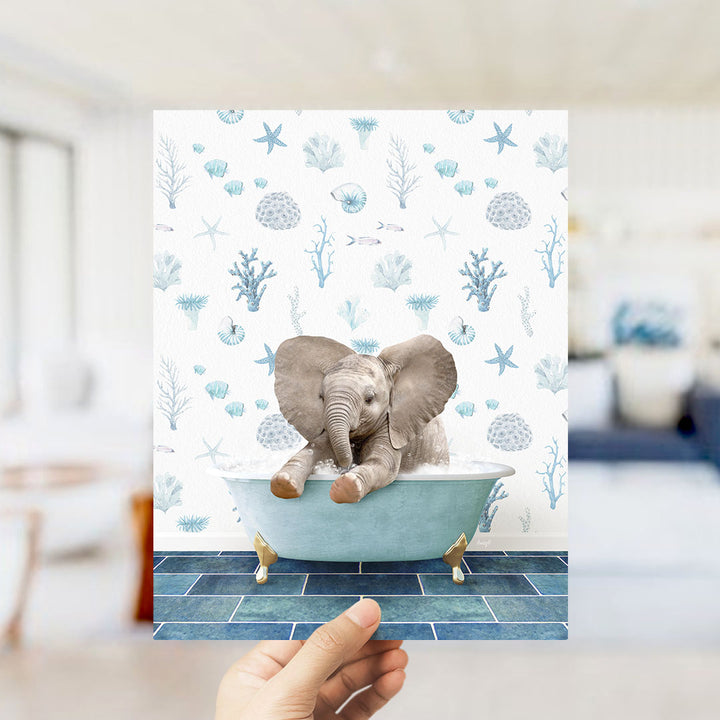 A hand is holding up a framed image of a baby elephant sitting in a bathtub filled with bubbles, surrounded by a blue and white ocean-themed wallpaper.