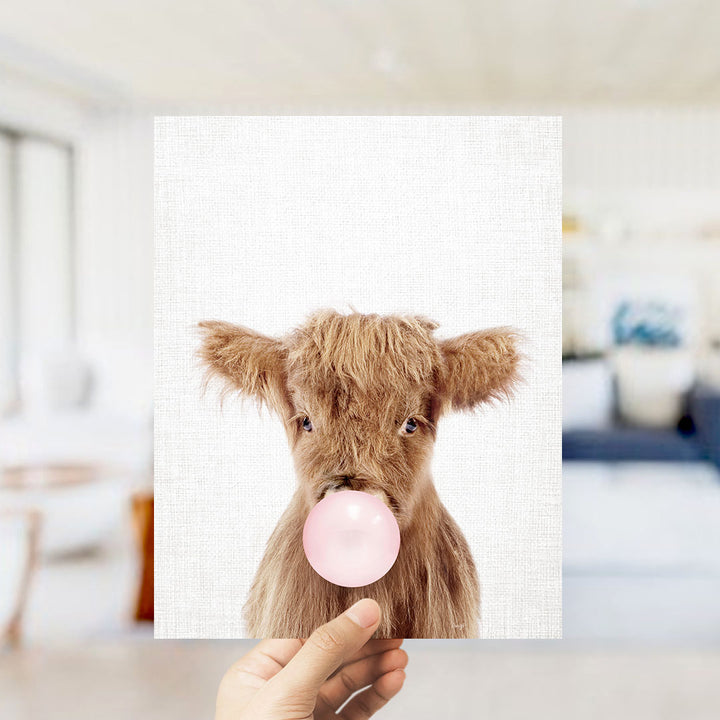 A hand is holding up a picture of a brown cow blowing a bubble with a pink bubble gum bubble.