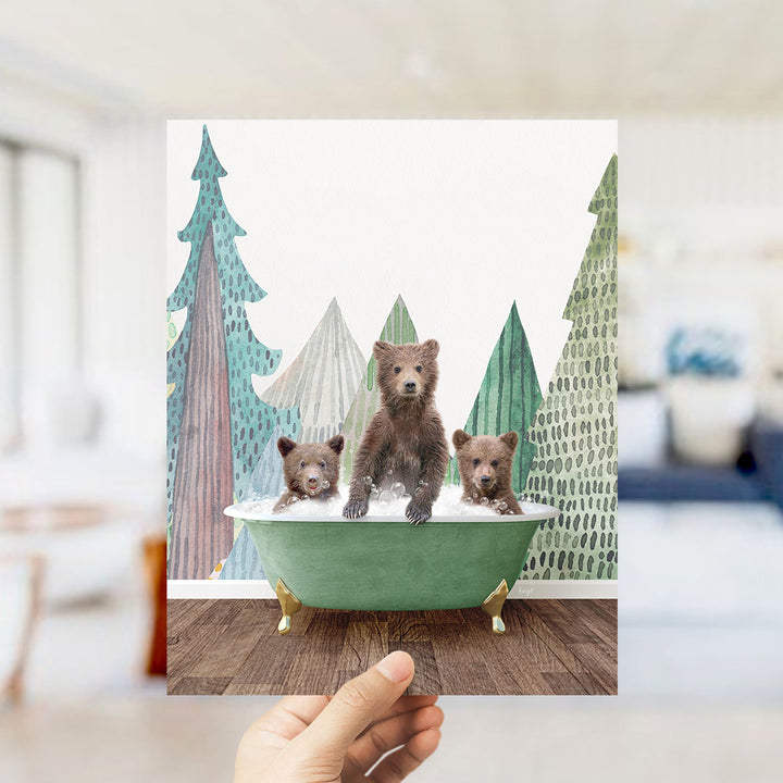A hand holds up a framed image depicting three bears sitting in a green bathtub, surrounded by a forest scene with trees and a snowy landscape.