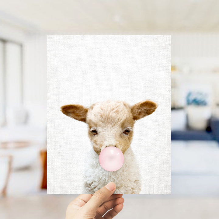 A hand is holding up a white piece of paper with a lamb's head blowing a pink bubble bubblegum bubble.