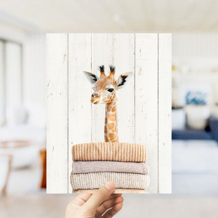A hand is holding up a stack of clothes, with a close-up view of a giraffe peeking out from behind the pile.