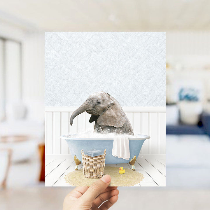 A hand holds up a framed image depicting a baby elephant sitting in a bathtub filled with water and bubbles, surrounded by a yellow rubber duck.