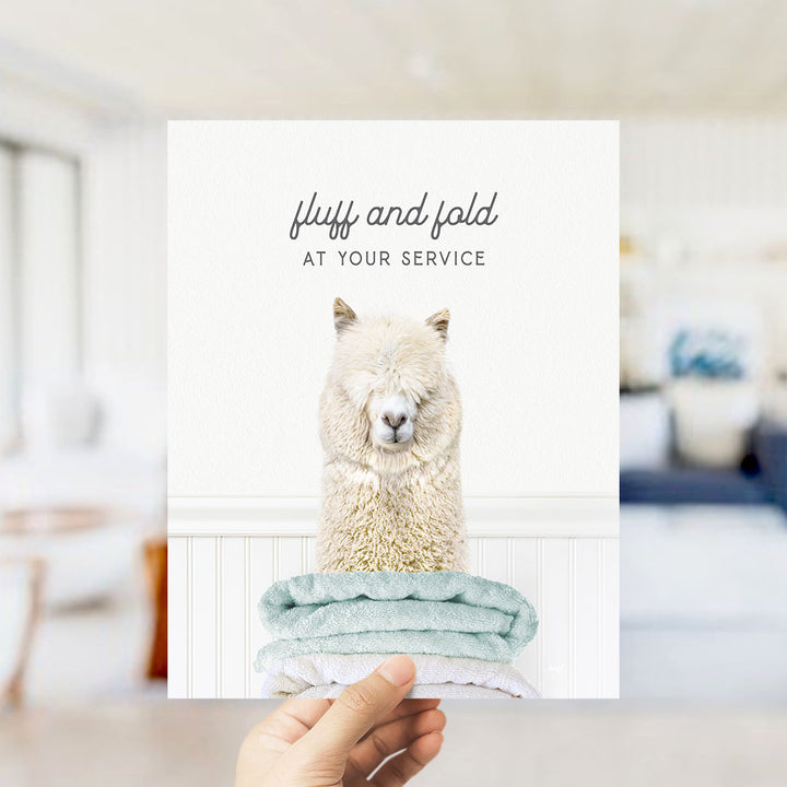 A hand holding a card with a photo of a fluffy white alpaca sitting on a stack of light blue towels, with the text "fluff and fold at your service" written above the image.
