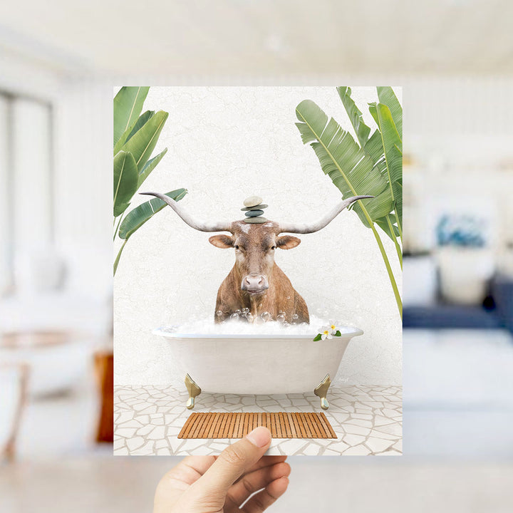 A hand holds up a framed image depicting a brown cow with large horns sitting in a white bathtub filled with bubbles, surrounded by tropical plants.