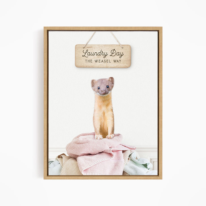A small brown cat sitting on a pile of clothes, with a wooden sign above that says "Laundry Day The Weasel Way".