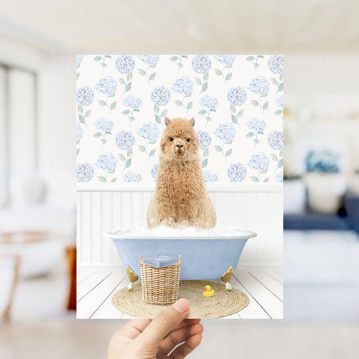 A hand holds up a framed image of a brown alpaca sitting in a blue bathtub filled with bubbles, surrounded by a floral patterned wall.
