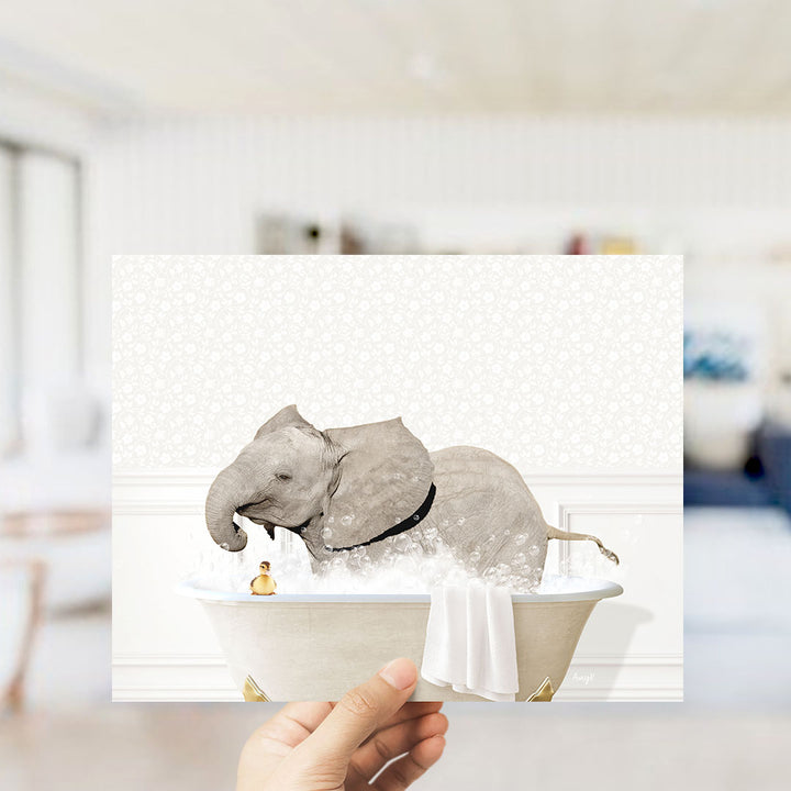 A hand holds up a framed image depicting an elephant taking a bath in a tub, with a small rubber duck floating nearby.