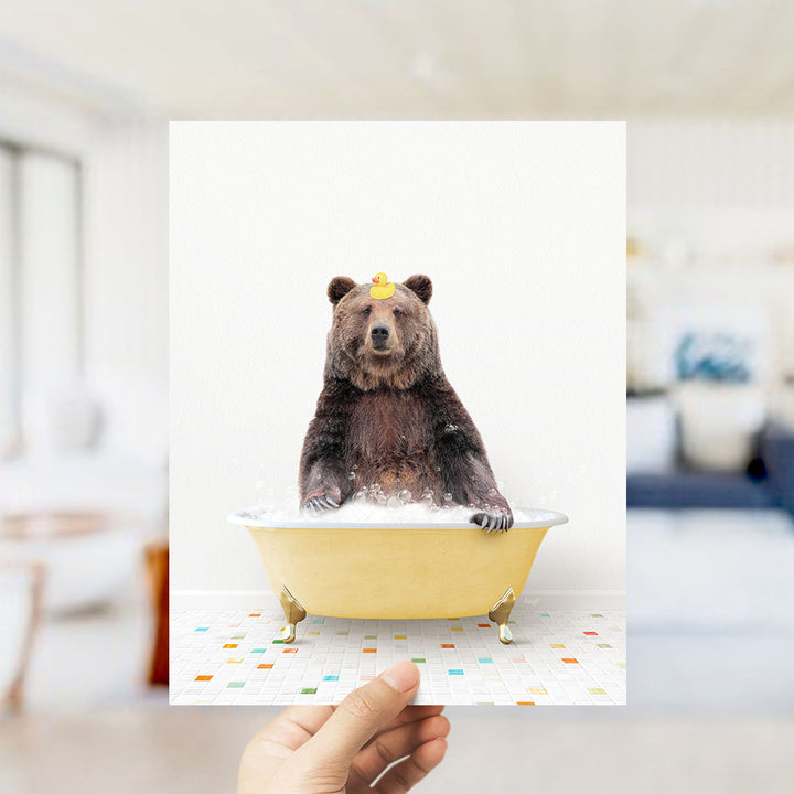 A hand holds up a framed image of a brown bear sitting in a yellow bathtub filled with bubbles, wearing a rubber duck hat.