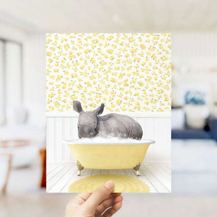 A hand holds up a framed image depicting a rhinoceros sitting in a yellow bathtub filled with bubbles, surrounded by a yellow and white floral pattern.