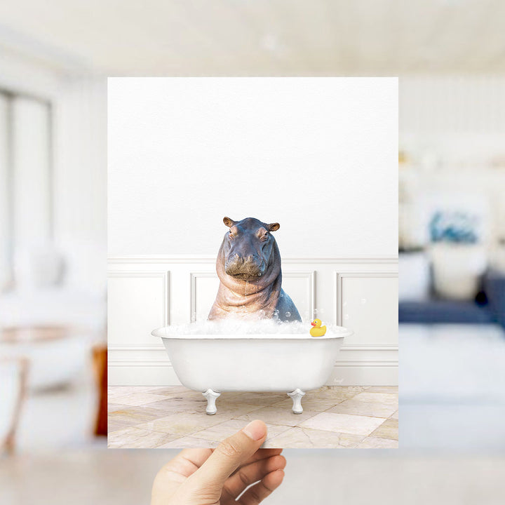 A hand holds up a framed image of a hippopotamus sitting in a bathtub, with a rubber duck floating in the water.