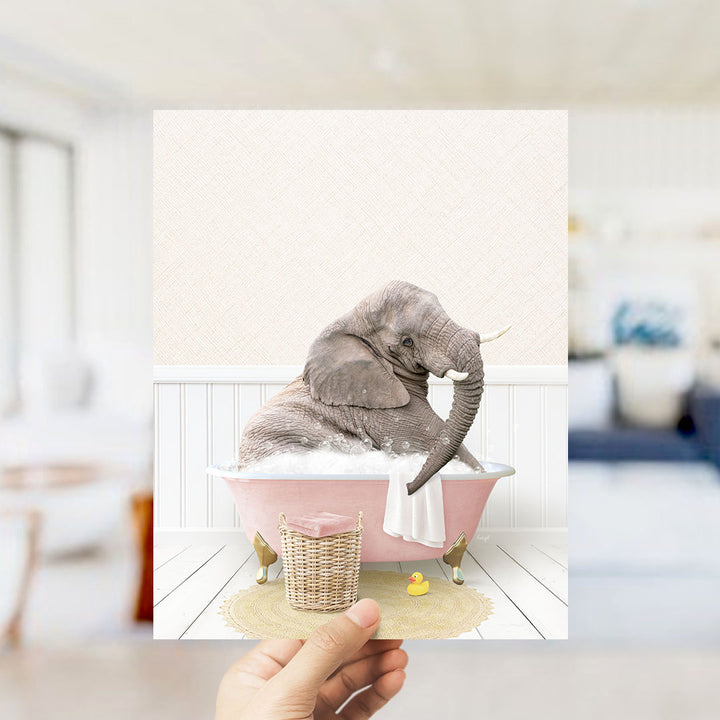 A hand holds up a framed image depicting an elephant sitting in a pink bathtub filled with bubbles, surrounded by a yellow rubber duck and a white towel.