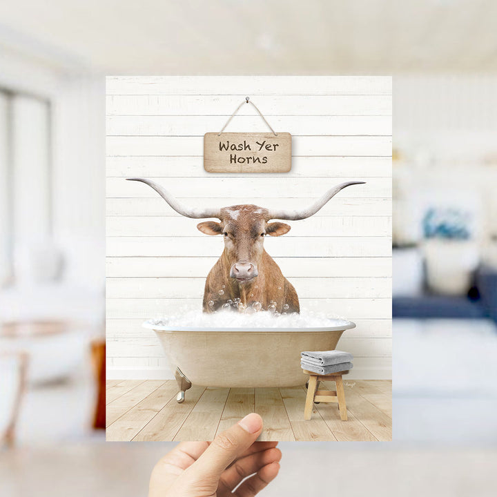 A hand holds up a framed image depicting a brown bull with long horns sitting in a bathtub, with a sign above that says "Wash Yer Horns".