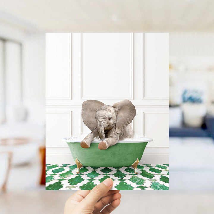 A hand holds up a framed image of a baby elephant sitting in a green bathtub filled with water, surrounded by a green and white patterned floor.