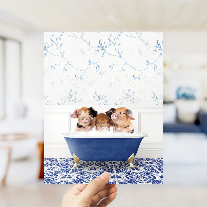 A hand holds up a framed image of three adorable pigs sitting in a blue bathtub, surrounded by a floral patterned wall.