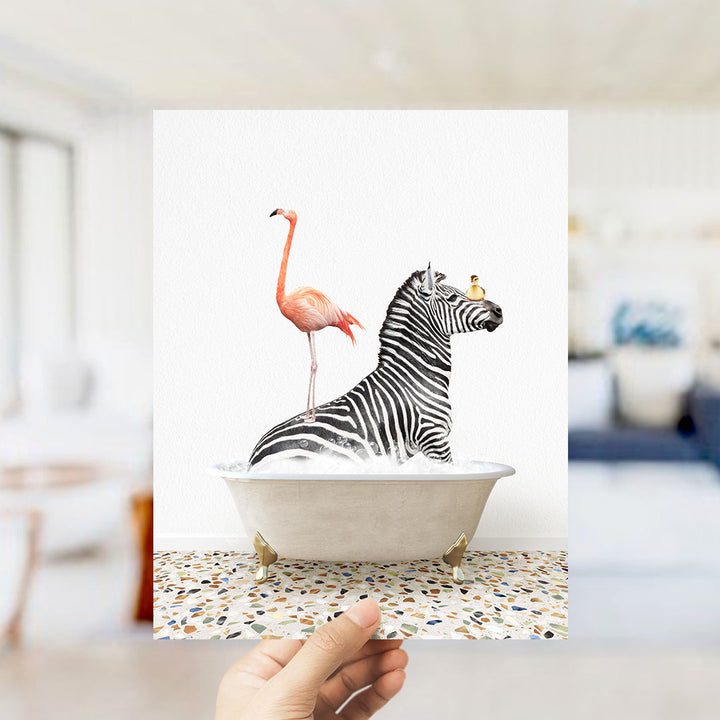 A hand holds up a framed image depicting a zebra standing on one leg inside a bathtub, with a pink flamingo standing on the zebra's back.