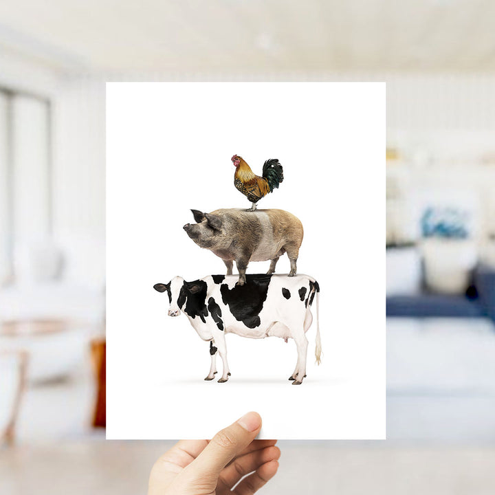 A hand is holding up a white card with an illustration of a cow, pig, and chicken standing on top of each other.