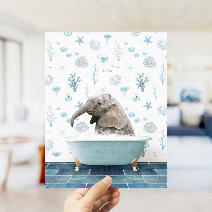 A hand is holding up a framed image of a baby elephant sitting in a bathtub filled with bubbles, surrounded by a blue and white ocean-themed wallpaper.
