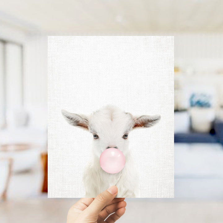 A hand is holding up a white goat with a pink bubble in its mouth, creating a playful and whimsical scene.
