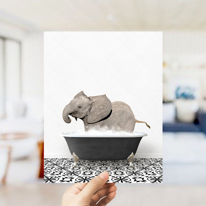 A hand holds up a framed image of a baby elephant sitting in a bathtub filled with water, surrounded by a black and white patterned floor.