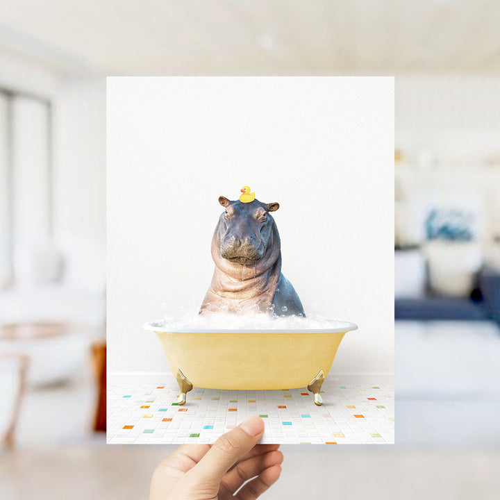 A hand holds up a framed image of a hippopotamus sitting in a bathtub, with a rubber duck perched on its head.