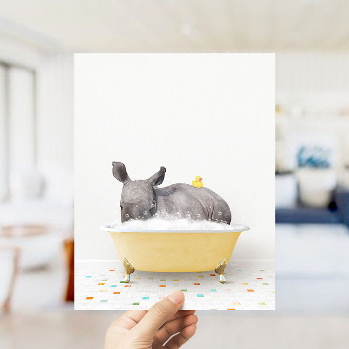 A hand holds up a framed image depicting a rhinoceros sitting in a yellow bathtub, with a yellow rubber duck floating on the water.