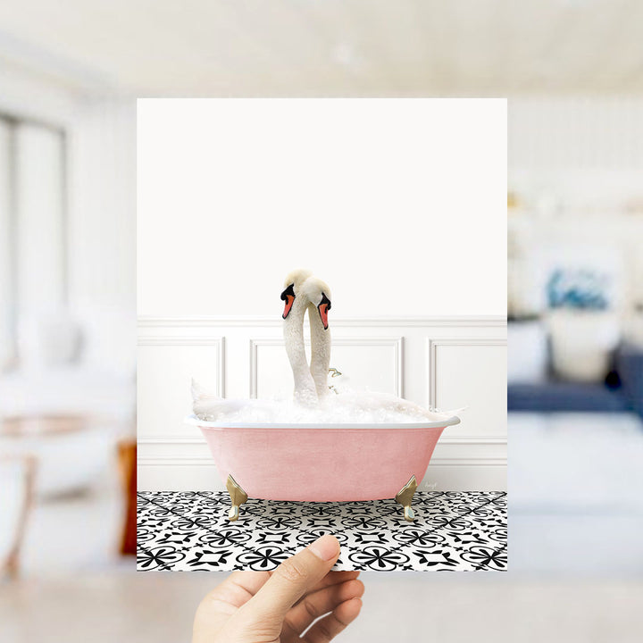 A hand holds up a framed image of two swans in a pink bathtub, with a black and white patterned floor in the background.