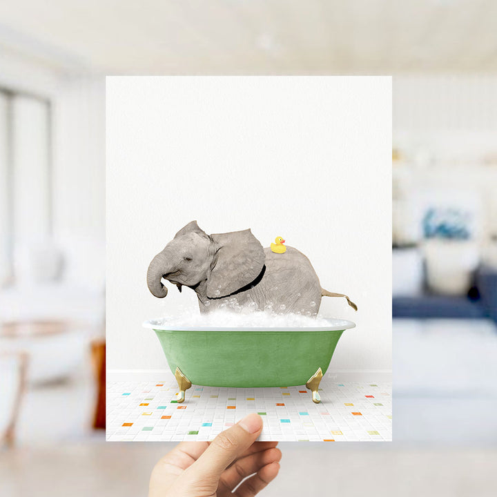 A hand holds up a framed image depicting a gray elephant sitting in a green bathtub, with a yellow rubber duck on its back.