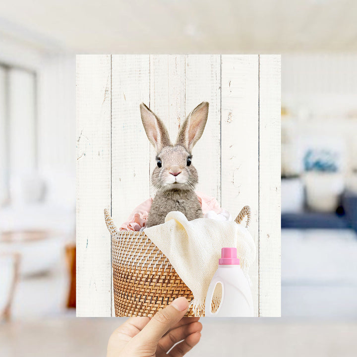 A small gray rabbit peeking out from a woven basket filled with a white cloth and a pink bottle.