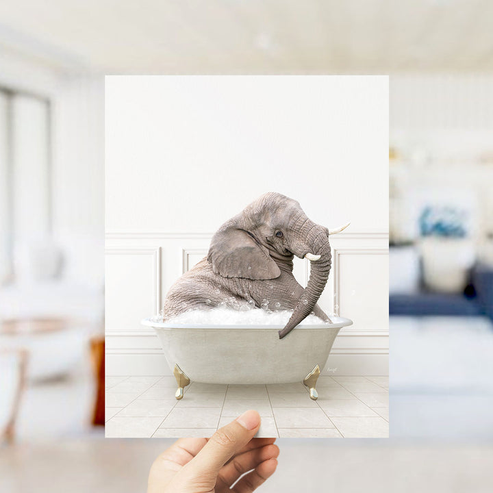 A hand holds up a framed image of a gray elephant sitting in a white bathtub filled with bubbles, creating a whimsical and playful scene.