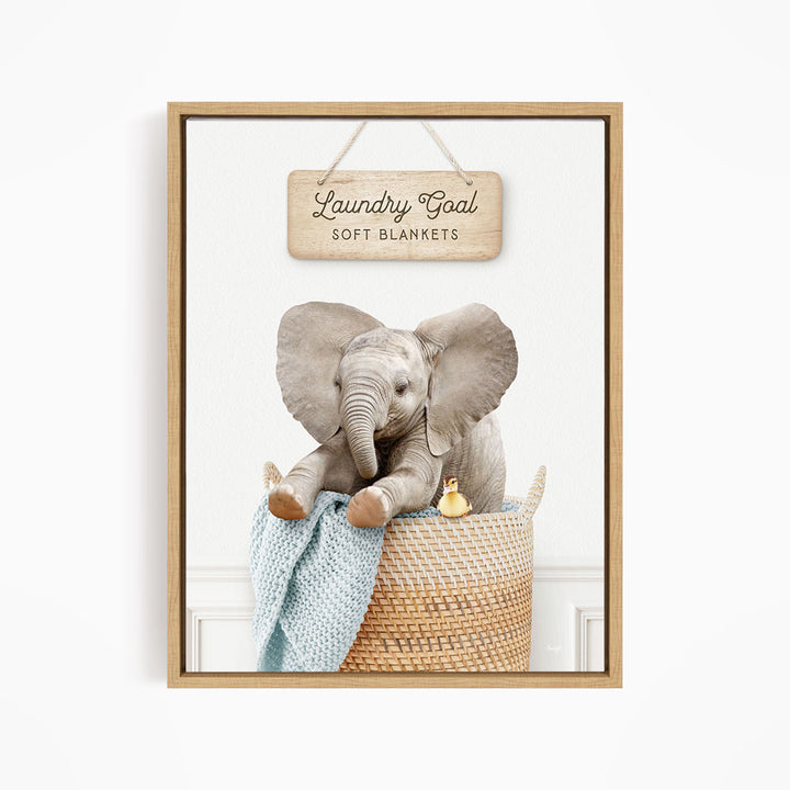 A framed image of an elephant sitting in a woven basket, with a sign above it that says "Laundry Goal Soft Blankets".