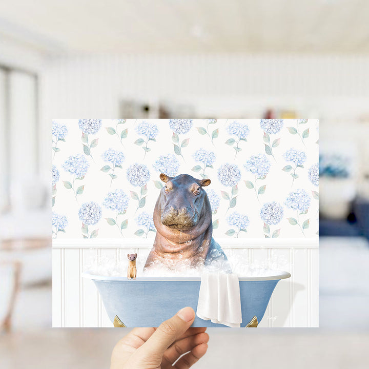 A hand holding a framed image of a hippopotamus sitting in a bathtub, surrounded by a floral pattern.