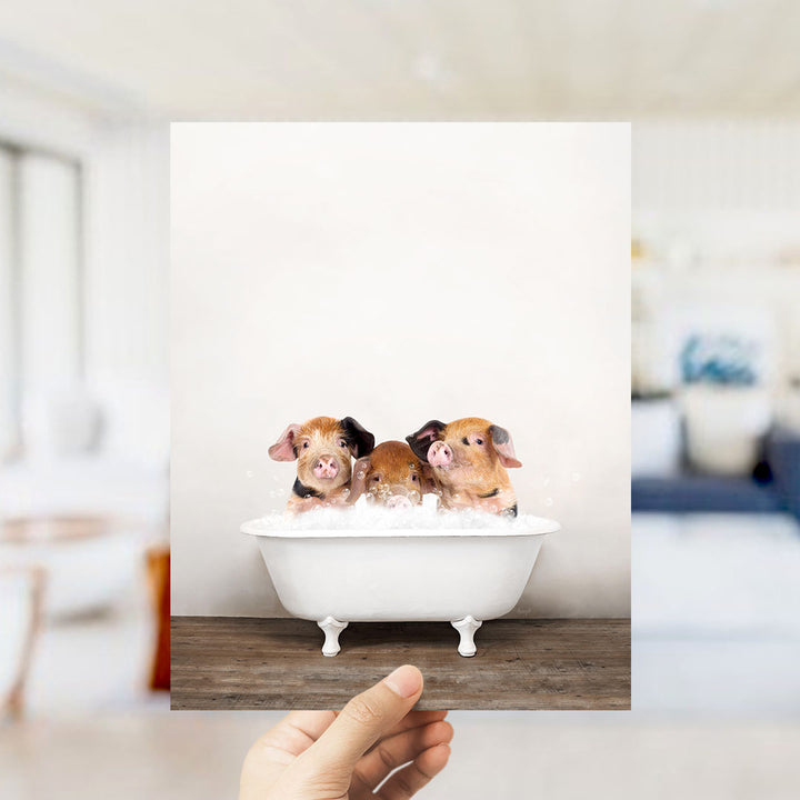 A hand holds up a framed photograph of three adorable pigs sitting in a white bathtub, with a blurred background suggesting a bathroom setting.