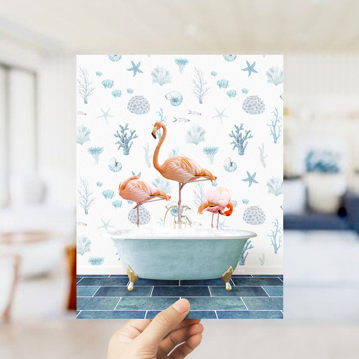 A hand is holding up a framed image of three flamingos sitting in a bathtub filled with bubbles, surrounded by a blue and white floral pattern.