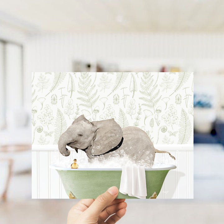A hand holds up a framed image depicting an elephant sitting in a bathtub, surrounded by a green bathtub, a small yellow duck, and a white towel.