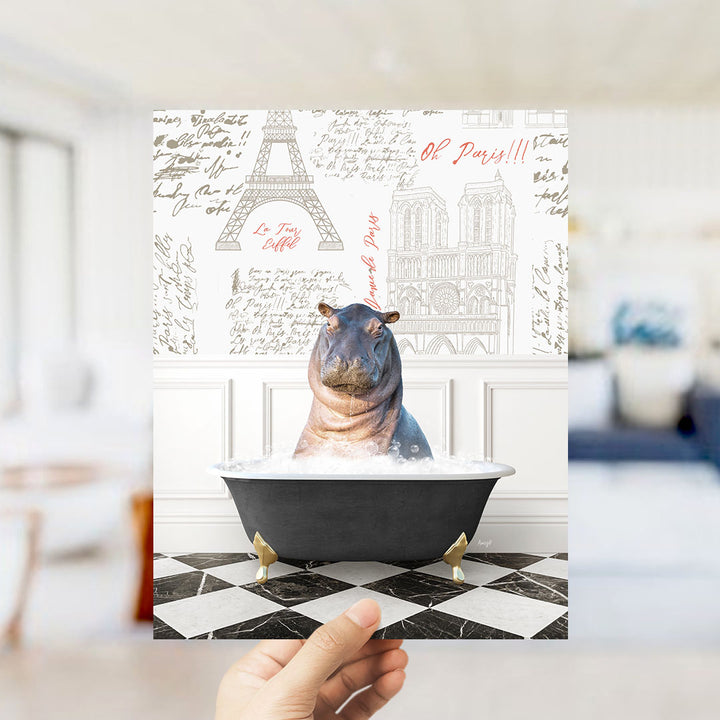 A hand is holding up a printed image featuring a hippopotamus sitting in a bathtub, surrounded by various Paris-themed elements such as the Eiffel Tower and the Notre-Dame Cathedral.