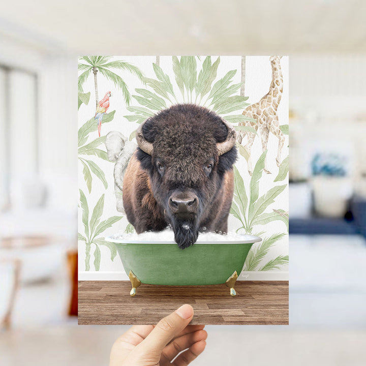A hand holds up a framed image depicting a bison in a bathtub, surrounded by tropical foliage.