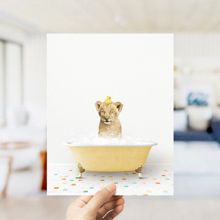 A hand holds up a framed image of a lion cub sitting in a yellow bathtub filled with bubbles, with a yellow rubber duck perched on its head.