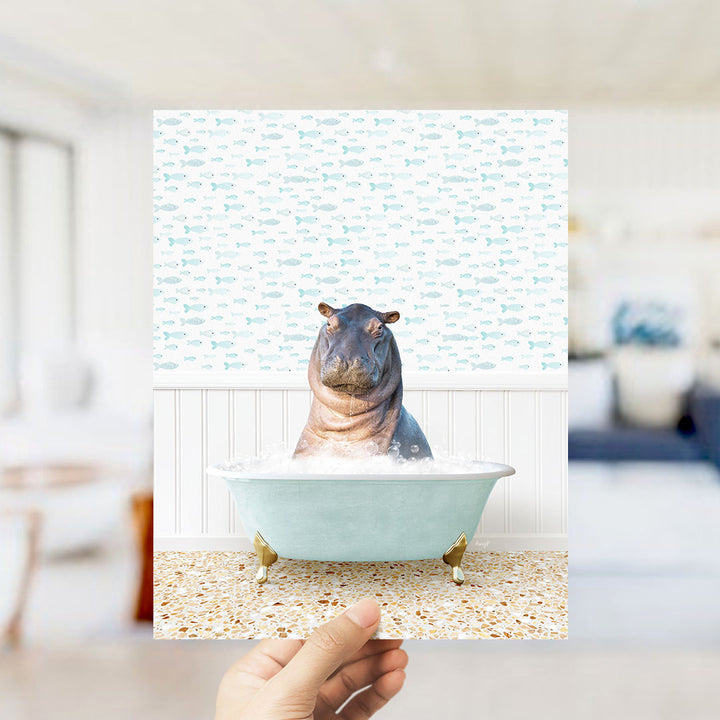 A hand holds up a framed image of a hippopotamus sitting in a bathtub filled with bubbles, surrounded by a pattern of small fish.