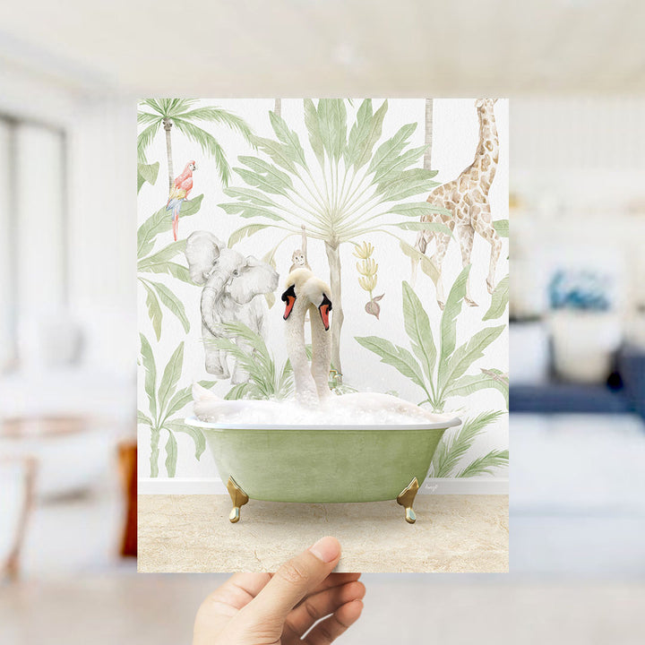 A hand holds up a framed print featuring a whimsical scene of two swans sitting in a bathtub surrounded by tropical foliage, including a giraffe and an elephant.